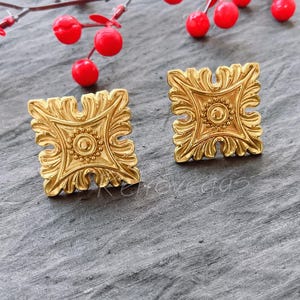 Może przedstawiać: Para złotych kwadratowych kolczyków z ozdobnym, szczegółowym wzorem. Kolczyki mają centralny okrągły wzór otoczony mniejszymi kółkami.