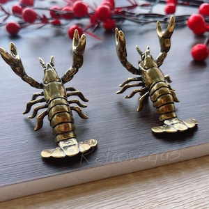 May include: Two brass-colored lobster figurines with raised claws and detailed bodies. The lobsters are positioned on a dark surface, with red berries and branches in the background. The figurines are approximately 4 inches long.
