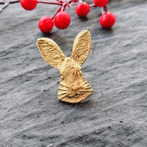 May include: A gold-colored rabbit head pin with detailed features. The pin is on a gray textured surface.