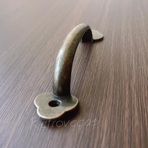 以下が含まれることがあります： 装飾的な花のデザインが施されたアンティークブラスのキャビネットプル。プルは湾曲した形状で、滑らかで経年変化した仕上げになっています。