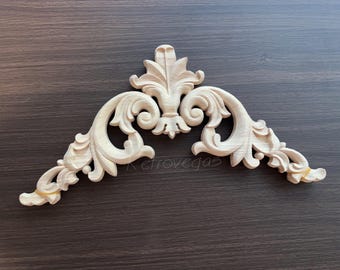 May include: A carved wooden decorative architectural element. The piece is light-colored, possibly unfinished, with a symmetrical design featuring scrolling leaves and floral details. The piece is likely for furniture or interior design.