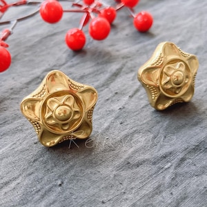 May include: A pair of gold-toned earrings with a star-shaped design. Each earring features a raised, circular centre with a smaller star design.