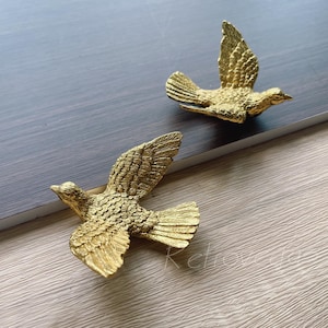 May include: Two gold-coloured bird-shaped decorative objects. The birds are in flight, with outstretched wings and detailed feather textures. They are placed on a dark grey and light brown surface.