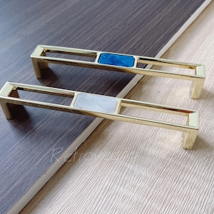 May include: Two gold cabinet door pulls with rectangular insets. One pull has a blue stone inset and the other has a white stone inset.
