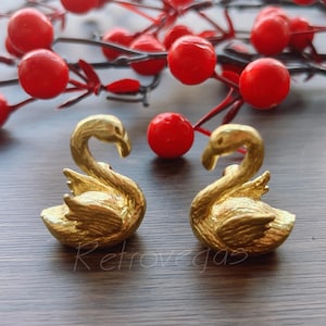 Può includere: Due statuette di cigni dorati si fronteggiano, con dettagliate texture di piume. Bacche rosse e rami sullo sfondo. La parola "Retrovegas" è visibile sulla superficie.