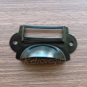 May include: Black metal drawer pull with a rectangular label holder. The pull has a rounded, slightly curved shape.