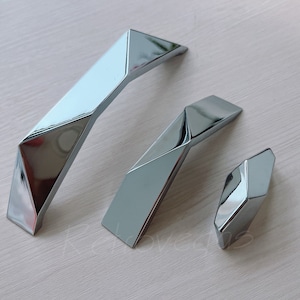 May include: Three silver-toned, geometric cabinet pulls in different sizes. The pulls have a modern, angular design.