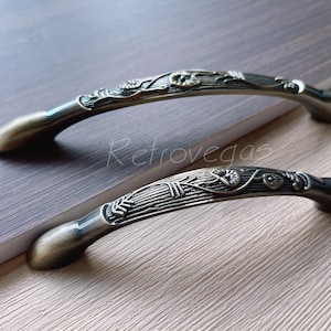 Könnte beinhalten: Zwei verzierte antike Schranktürgriffe mit floralen Designs. Die Griffe sind aus Metall und haben eine Bronze-Oberfläche.