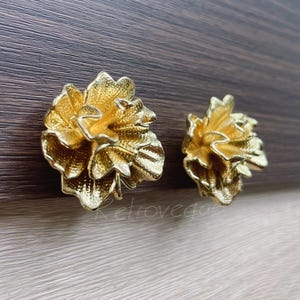 May include: Two gold-colored flower-shaped earrings. The earrings have detailed petal designs and a shiny finish. The earrings are set against a dark wood-grain background. The earrings are likely vintage or costume jewelry.