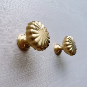 Op de afbeelding: Twee gouden kastknoppen met een geribbeld, zonnestralenontwerp. De knoppen zijn gemonteerd op een wit oppervlak.