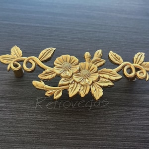 May include: Gold-toned metal drawer pull with a floral design. The pull features two stylized flowers with leaves and vines.