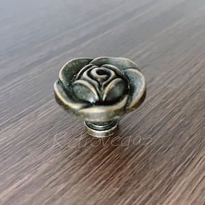 Op de afbeelding: Een bronzen kastknop met een rozenontwerp. De knop heeft gedetailleerde bloemblaadjes en een ronde basis. De hardware is geplaatst op een donker houten oppervlak. Het woord "Retrovegas" is vaag zichtbaar.