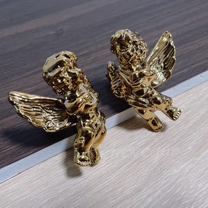 Peut inclure: Deux figurines de chérubins dorés avec des ailes, l'une tenant une trompette et l'autre une fleur.