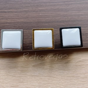 Op de afbeelding: Drie vierkante kastknoppen met een witte keramische inzet. De knoppen zijn in zilver, goud en zwart.