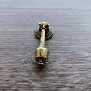 May include: A vintage-style, antique brass drawer pull with a decorative design. The pull has a round base and a long, slender handle.