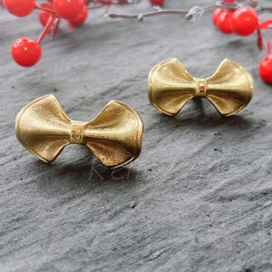 May include: A pair of gold bow-shaped earrings. The earrings are made of a smooth, shiny metal and have a simple, elegant design.