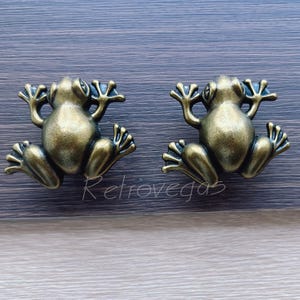 Op de afbeelding: Twee bronzen klinken in de vorm van een kikker. De klinken hebben gedetailleerde uitgestrekte ledematen en een getextureerde afwerking. De hardware is gemonteerd op een houten oppervlak. Het woord "retrovegas" staat onderaan in cursief.