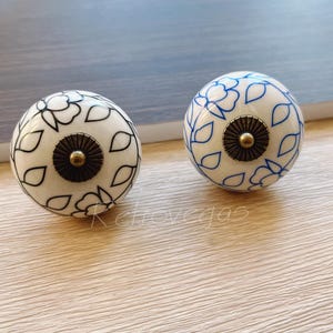 May include: Two white ceramic cabinet knobs with blue and black floral designs. The knobs have a gold metal accent in the center.
