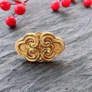 May include: A gold-coloured metal ring with a detailed design of swirling clouds. The ring is on a grey textured surface.