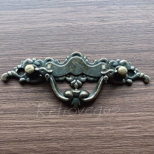 May include: An ornate antique-style drawer pull with a decorative design and two round knobs. The pull is made of metal and has a dark bronze finish.