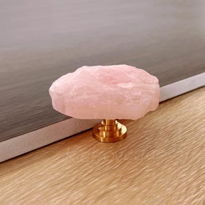 Op de afbeelding: Een rozenkwarts kastknop met een goudkleurige basis. De roze steen heeft een ruwe, natuurlijke textuur. De knop staat tegen een neutrale achtergrond.
