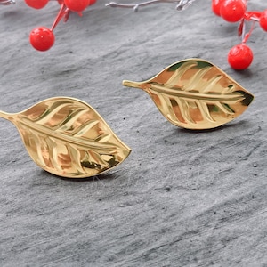 Puede incluir: Un par de pendientes de hoja de oro. Los pendientes están hechos de un metal brillante y tienen un diseño de hoja detallado.