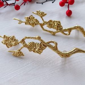 Op de afbeelding: Twee goudkleurige metalen decoratieve stukken in de vorm van gestileerde takken met delicate bloemen. De stukken zijn op een wit oppervlak gerangschikt.