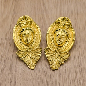 Könnte beinhalten: Ein Paar goldfarbene Ohrringe mit einem detaillierten Design, das ein Frauen Gesicht in einem dekorativen Rahmen zeigt.