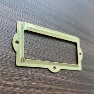 May include: A gold-coloured metal label holder with a rectangular opening for inserting a label. The holder has a decorative design around the edges.