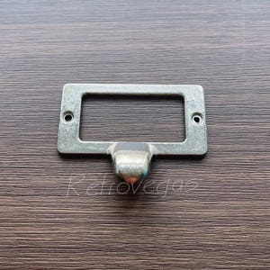 Puede incluir: Un tirador de cajón de metal plateado con una placa rectangular y un asa redondeada. El tirador tiene un acabado ligeramente envejecido.