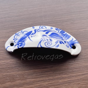 Puede incluir: Tirador de cajón de cerámica blanca con un diseño floral azul y blanco. El tirador tiene forma de semicírculo.