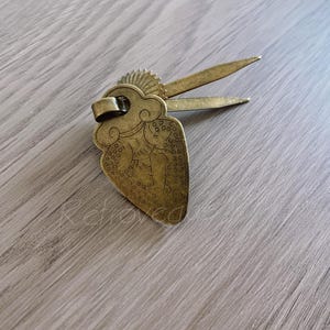 May include: A gold-colored metal hair pick with a heart-shaped design. The heart has a detailed design of a person with a cloud above their head. The pick has a long, thin handle.