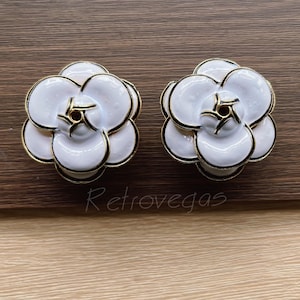 May include: Two white flower-shaped decorative objects with gold trim and centres. The objects are on a wooden surface. The text "Retrovegas" is visible in the image.