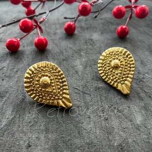 Könnte beinhalten: Ein Paar goldfarbene, tropfenförmige Ohrringe mit einem strukturierten Design. Die Ohrringe haben einen kleinen Kreis in der Mitte jedes Tropfens.