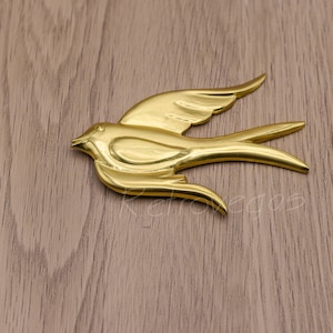 Puede incluir: Una figurita de pájaro de metal de color dorado con un diseño estilizado. El pájaro está en vuelo con las alas extendidas y las plumas de la cola extendidas.