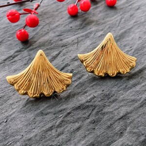 May include: A pair of gold-tone earrings shaped like ginkgo leaves. The earrings are on a gray textured surface.