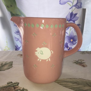 May include: A pink ceramic pitcher with a green and white design featuring a pig. The pitcher has a handle and a spout.