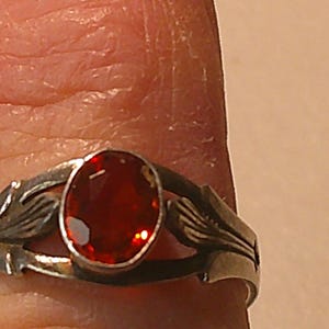 May include: A silver ring featuring an oval, deep red gemstone. The ring's band has a decorative leaf design on each side of the gemstone. The ring is shown on a finger.