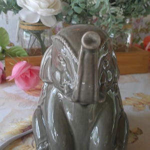 May include: A gray ceramic elephant figurine with a raised trunk and detailed carvings. The elephant is sitting, with its trunk pointing upwards. The figurine is on a floral patterned surface, with flowers and greenery in the background.