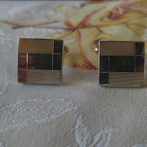 May include: A pair of gold-toned cufflinks with a geometric design. Each square cufflink features a combination of horizontal lines and rectangular insets. The cufflinks are set against a patterned background.