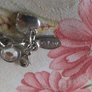 Puede incluir: Pulsera de abalorios plateada con abalorios de setas rosas y blancas, un abalorio de cristal transparente y una etiqueta DKNY. La pulsera est&aacute; sobre un fondo floral con flores rosas.