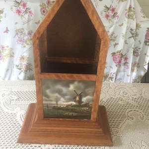 May include: A wooden house-shaped shelf with a small shelf inside and a picture of a windmill on the bottom shelf.