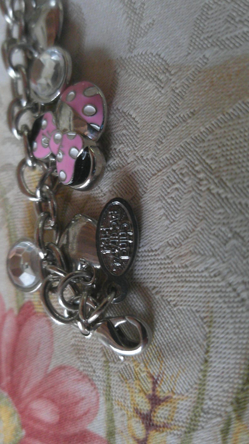 Puede incluir: Pulsera de abalorios plateada con un dije de hongo rosa y blanco, detalles de cristal transparente y un cierre de mosquet&oacute;n. La pulsera tambi&eacute;n presenta un peque&ntilde;o dije ovalado con texto.