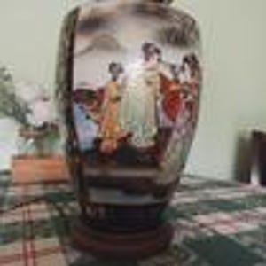May include: A decorative ceramic vase with a detailed painted scene of figures in traditional attire. The vase features a dark base and a wooden stand. The artwork includes shades of gold, red, and green, set against a landscape background.