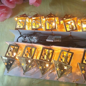 May include: A string of ten gold-colored metal lantern lights, each featuring star-shaped cutouts. The lanterns are arranged in two rows on a white cardboard display, with a black electrical cord connecting them and glowing warm yellow light.