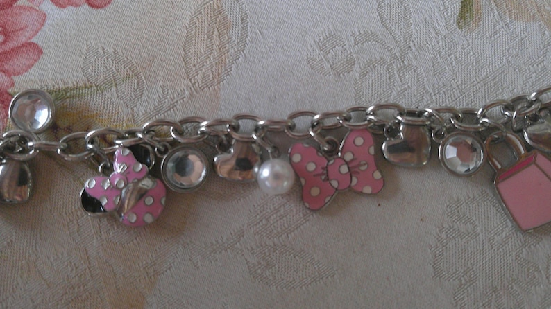 Puede incluir: Pulsera de abalorios plateada con varios abalorios, incluyendo un lazo rosa con lunares blancos, una mariquita, una perla, corazones y un bolso rosa. La pulsera tambi&eacute;n presenta abalorios transparentes, redondos y en forma de l&aacute;grima.