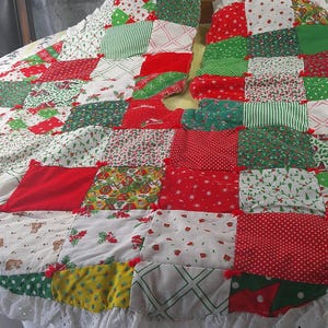 May include: A circular Christmas tree skirt featuring a patchwork design of various red, green, and white fabric squares. The quilt-style textile includes patterns of holly, stripes, and dots, finished with a white eyelet lace trim along the edge.