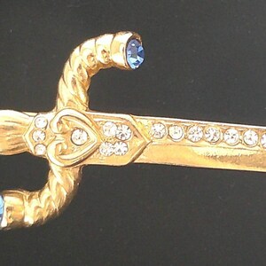 May include: A gold-toned brooch in the shape of a dagger with a heart-shaped design. The dagger is decorated with clear rhinestones and has red, green, and blue gemstones.