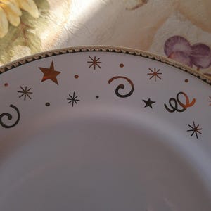 May include: White ceramic plate with a gold rim and festive star and swirl designs. The plate features copper-colored stars, swirls, and dots, creating a celebratory aesthetic. The plate is suitable for serving food.