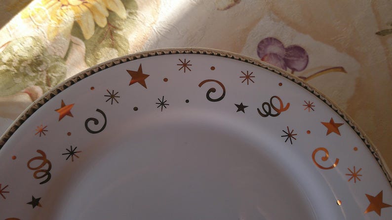 May include: White ceramic plate with a gold rim and festive star and swirl designs. The plate features copper-colored stars, swirls, and dots, creating a celebratory aesthetic. The plate is suitable for serving food.